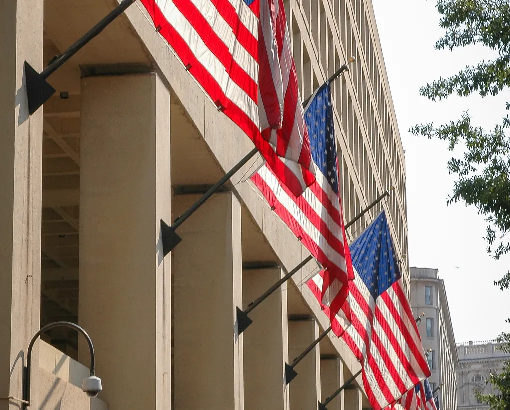 The image shows American flags hanging from a beige building, mounted on black brackets along the columns, daytime.