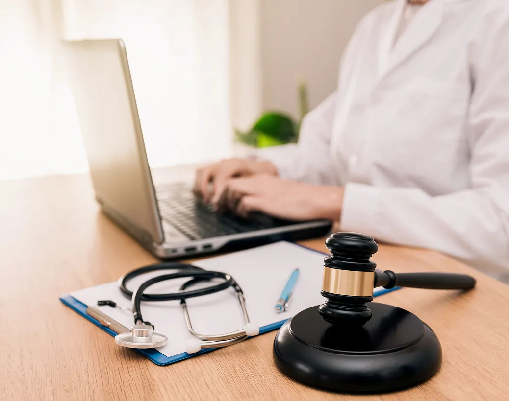 A doctor works on a laptop at a desk with a clipboard, stethoscope, and a judge’s gavel, implying a medical-legal context.