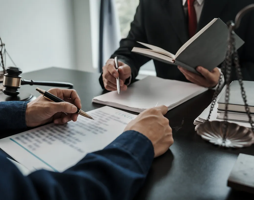 Two people sign a document in a legal setting, with a gavel, Scales of Justice, and a book nearby.