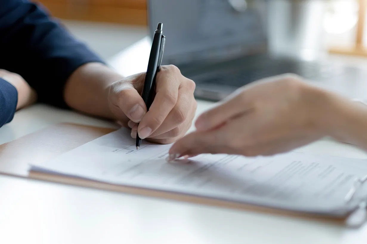 Two hands sign a document on a clipboard with a pen, a laptop blurred in the background.