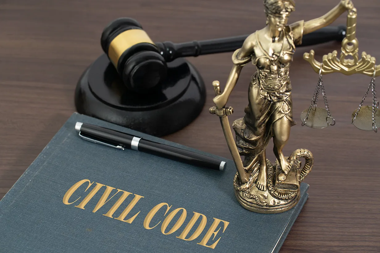 Gold statue of Lady Justice with scales and sword, a gavel, and a navy blue "CIVIL CODE" book with a pen on a wooden desk.