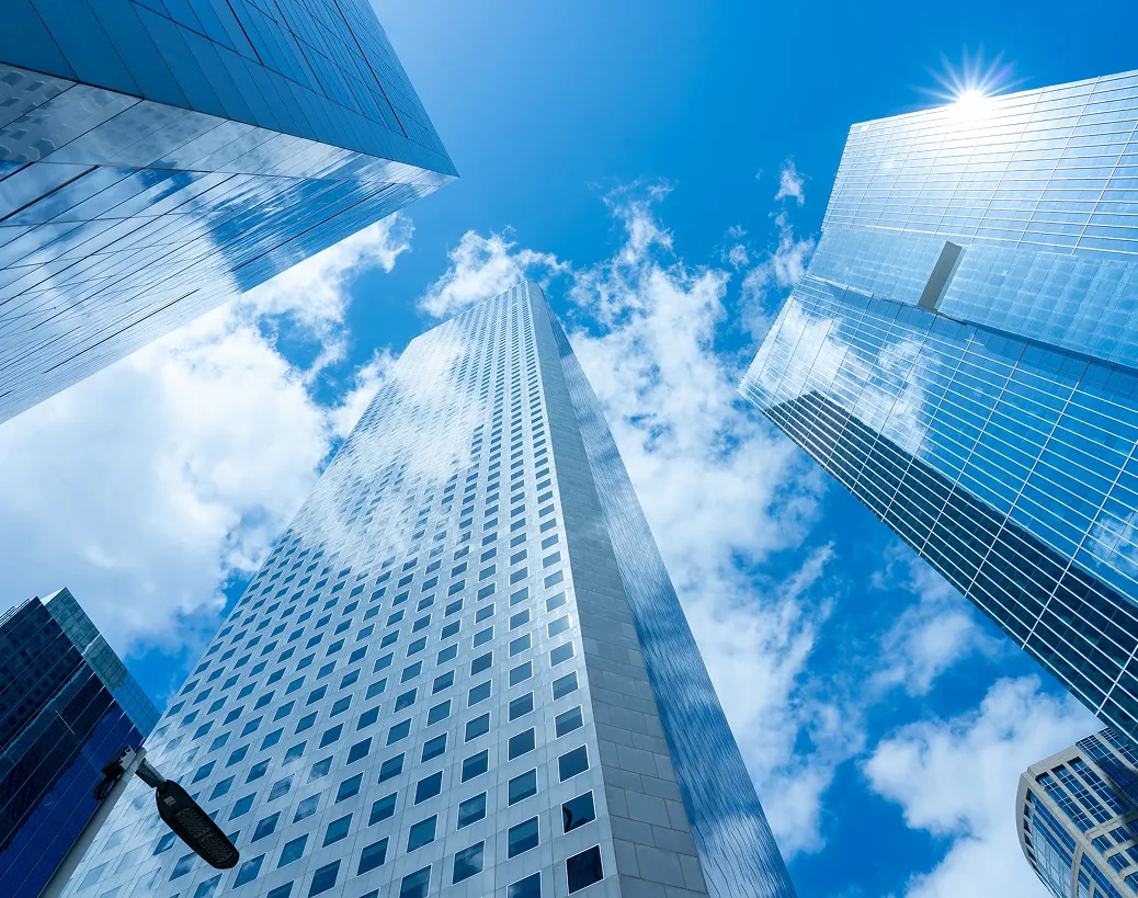 Several tall, reflective glass skyscrapers rise against a bright blue sky with clouds, capturing sunlight off the upper edge of the tallest building.
